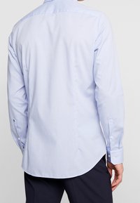 Jasnoniebieska koszula z długim rękawem wykonana z gładkiej tkaniny, z klasycznym kołnierzem, zaokrąglonym dołem i mankietami zapinanymi na guziki. Widoczne subtelne pionowe paski.