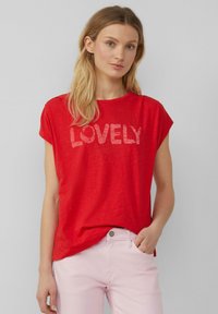 T-shirt rouge à manches courtes arborant le mot "LOVELY" en rouge clair. Texture douce avec une coupe décontractée, associé à un pantalon rose clair.