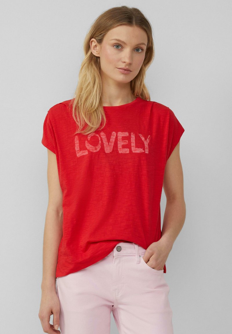 T-shirt rouge à manches courtes arborant le mot "LOVELY" en rouge clair. Texture douce avec une coupe décontractée, associé à un pantalon rose clair.