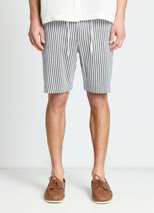 Uomo che indossa pantaloncini a righe verticali grigie e bianche con coulisse e scarpe da barca in pelle marrone, in piedi su un pavimento bianco davanti a uno sfondo semplice.