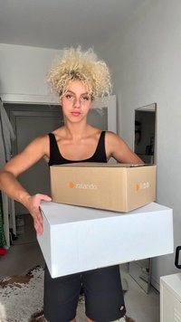 Jsou zde dvě kartonové krabice: jedna hnědá s logem Zalando a jedna bílá. V pozadí se nachází zrcadlo a světlé stěny.