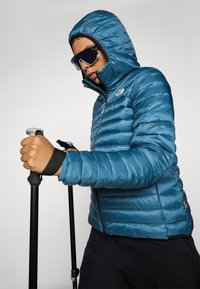 Teal pufferjack met een capuchon, met een horizontaal gestikt ontwerp, ritssluiting en logo. Model houdt trekkingstokken vast en draagt een zonnebril.