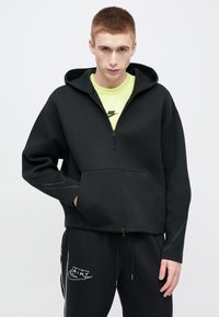 Μαύρο φούτερ με φερμουάρ και μεγάλο κουκούλι, κατασκευασμένο από απαλό ύφασμα. Διαθέτει εμπρόσθια τσέπη και ραφές σε αντίθεση. Φοριέται πάνω από ένα νέον πράσινο πουκάμισο.