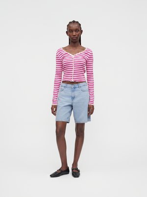Kvinde med fletninger iført en pink og hvid stribet cropped cardigan, lyse denim Bermuda-shorts og sorte ballerinaer, står foran en hvid baggrund.