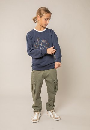 Garçon ajustant la manche d'un sweat-shirt bleu marine avec un motif de trottinette, portant un pantalon cargo olive et des baskets blanches sur fond neutre.