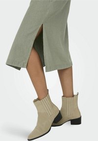 Stivaletti beige in suede con punta affusolata, dettagli a coste e tacco basso nero, abbinati a una gonna verde con spacchi laterali.