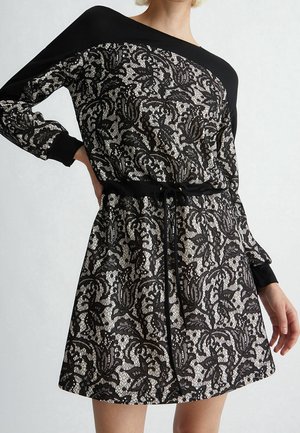 Kobieta ubrana w czarną i beżową koronkową sukienkę w kwiatowy wzór z długimi rękawami oraz czarnym paskiem ze ściągaczem w talii na jednolitym tle.