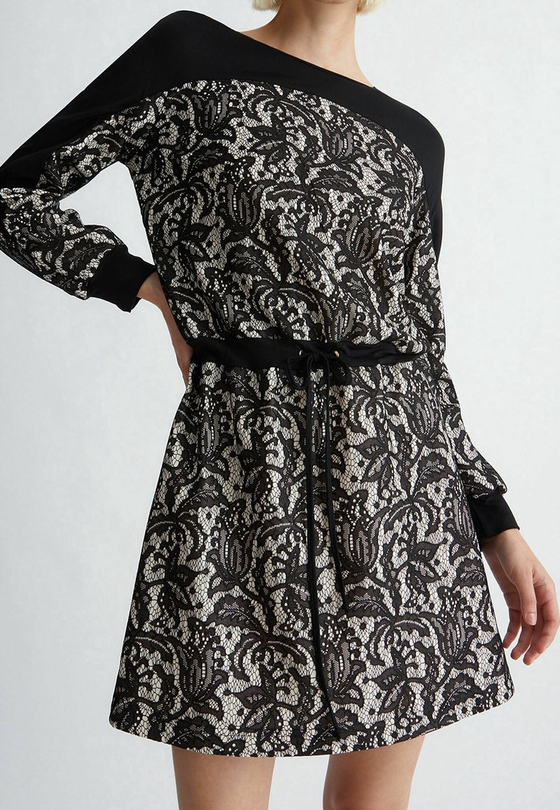 Femme portant une robe à manches longues noire et beige à motifs floraux en dentelle, avec une ceinture à cordon noire à la taille, sur un fond uni.