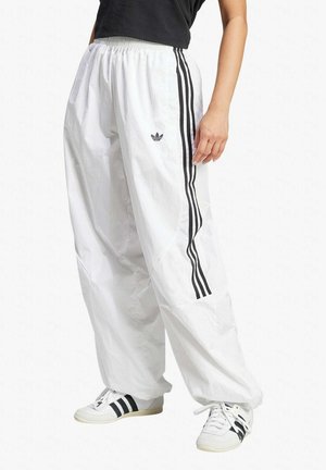 Henkilö pukeutuneena löysiin valkoisiin Adidas-verkkarihousuihin mustilla raidoilla ja valkoiset lenkkarit mustilla raidoilla, seisoo valkoista taustaa vasten.