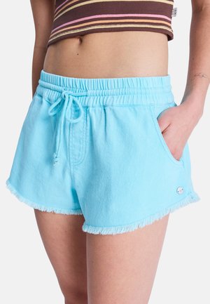 Kobieta w jasnoniebieskich elastycznych szortach z sznurkiem i surowym obszyciem, z ręką w kieszeni, oraz w brązowym, paskowanym, krótko przyciętym topie.