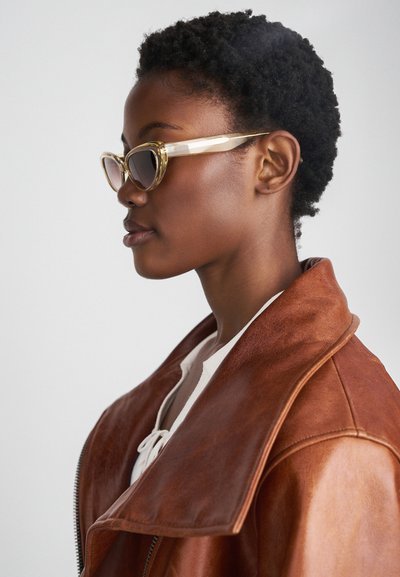 Chaqueta de cuero marrón con un gran cuello; gafas de sol estilo gato en tonos claros y marrones; cabello negro corto y texturizado; fondo neutro.