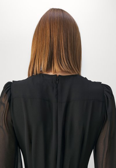 Robe noire avec des manches bouffantes transparentes, présentant un col haut et une fermeture éclair discrète au dos. Texture lisse, design minimal et coupe ajustée.