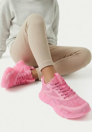 Dítě sedící se zkříženýma nohama, oblečené v béžových žebrovaných legínách a jasně růžových síťových teniskách Reebok s tlustou podrážkou.