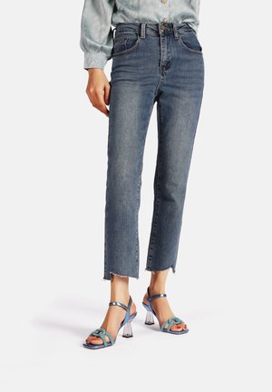 Jeans in denim blu a vita alta con orlo grezzo, abbinati a sandali blu con cinturini e tacchi a blocco trasparenti con dettagli a nodo.
