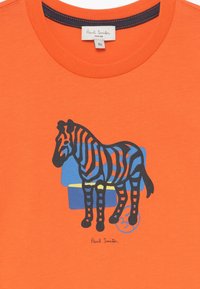 Camiseta de algodón naranja con un gráfico de una cebra en negro y azul. Incluye un detalle de firma "Paul Smith" en la parte inferior.