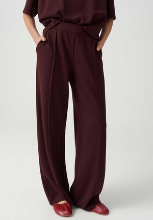 Persona che indossa pantaloni larghi bordeaux scuro con le mani nelle tasche e scarpe basse rosse abbinate su uno sfondo chiaro.