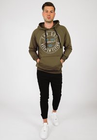 Olivgrön hoodie med en cirkulär grafik, "TOP GUN" i vitt och blått, kombinerad med svarta slim-fit byxor och vita sneakers.