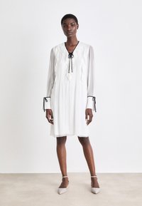 Weißes, knielanges Kleid mit langen, durchsichtigen Ärmeln, Rüschendetails und einer schwarzen Krawattenverschluss am Ausschnitt. Verfügt über schmale Manschetten mit Bändern.