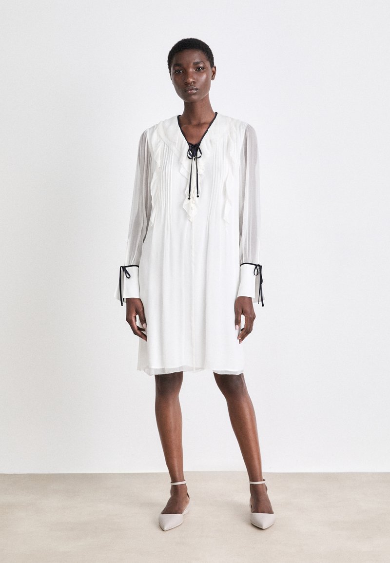 Weißes, knielanges Kleid mit langen, durchsichtigen Ärmeln, Rüschendetails und einer schwarzen Krawattenverschluss am Ausschnitt. Verfügt über schmale Manschetten mit Bändern.