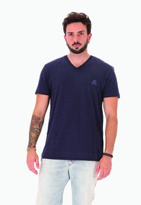 Uomo con il braccio sinistro tatuato e braccialetti di perline, indossa una t-shirt a V blu navy di Diesel e jeans azzurri, in piedi.