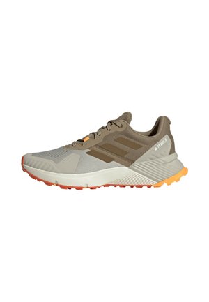 Zapatilla de trail running Adidas Terrex beige y marrón con suela naranja y entresuela blanca, vista lateral sobre fondo blanco.