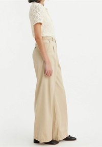 Kvinna klädd i en krämfärgad kortärmad stickad topp, högmidjade vida beige byxor och svarta låga klackskor, stående på sidan.