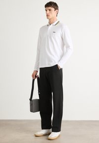Hvit langermet polo med en gul og grønn stripet krage, med en liten logo. Sammen med sorte bukser og hvite sneakers.