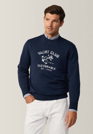 Felpa navy con grafica bianca "Yacht Club", collo a giro, polsini e orlo a coste. Indossata sopra una camicia a motivi quadrati blu chiaro e pantaloni bianchi.