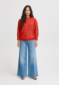 Pull en laine rouge avec un motif en torsades, associé à un jean en denim bleu clair à jambes larges. Chaussures en cuir noir à semelles plates.
