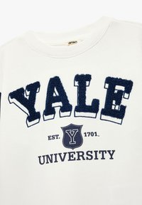 Biała bluza z haftowanym granatowym napisem "YALE" oraz wzorem herbu, z datą założenia "EST. 1701" i napisem "UNIWERSYTET" poniżej.