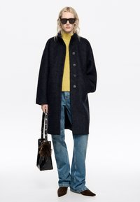 Jūras spēku zilo vilnas maisījuma mētelis ar melniem pogām, valkāts virs dzeltena džempera. Apvienots ar ziliem džinsiem, melnu somu ar kažokādas detalizāciju un brūnām kurpēm.