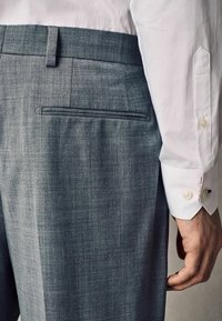Pantalones grises ajustados con una tela texturizada, que presentan un frente plano y un bolsillo trasero. La camisa a juego es blanca con puños abotonados.