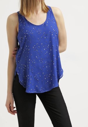 Kvinden bærer en ærmeløs blå top med små hjerte mønstre, kombineret med sorte bukser, stående mod en ensfarvet hvid baggrund.