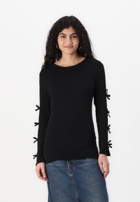 Schwarzes Langarmoberteil mit rundem Halsausschnitt, verziert mit Schleifen an den Ärmeln. Das Material wirkt glatt und figurnah. Kombiniert mit einem Jeansrock.