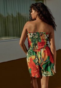 Trägerloser Jumpsuit mit gerafftem Oberteil, mit einem bunten tropischen Blumenmuster, das Grün-, Rot- und Gelbtöne kombiniert, aus leichtem Stoff gefertigt.