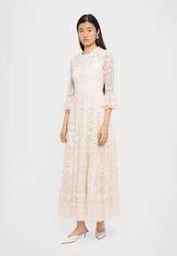 Lange, blush-kleurige jurk met bloemenstickers, doorschijnende mouwen en een uitlopende rok. Bevat gerimpelde manchetjes en een aansluitend lijfje.