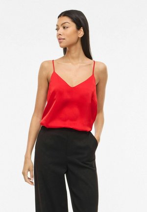 Kvinde med langt mørkt hår iført en rød ærmeløs top og sorte bukser med høj talje, stående med den ene hånd i lommen og kiggende mod venstre.