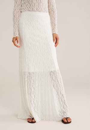 Naine, kes kannab pikka valget pitsseelikut ja sobivat pitsist tappi, seisab mustades sandaalides neutraalsel taustal.