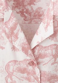 Chemise à col avec motif toile rose mettant en scène des tigres et des arbres sur fond blanc, fermée par un unique bouton rose.