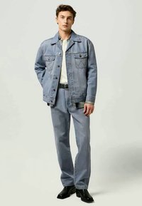 Jaqueta e calças de ganga em azul claro, com quatro bolsos frontais, fechos de botão e costuras contrastantes. O modelo está de pé com um fundo neutro.