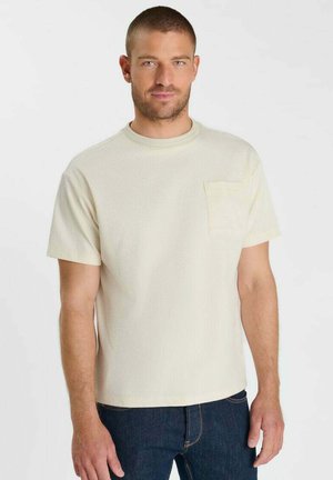 Man met kort haar, die een lichtbeige gestructureerd shirt met korte mouwen en een borstzak draagt en een donkerblauwe spijkerbroek, staand tegen een witte achtergrond.