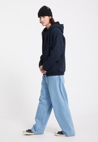 Jeune homme marchant, portant un bonnet noir, un sweat à capuche bleu marine, un jean large bleu clair et des baskets noires sur fond blanc uni.