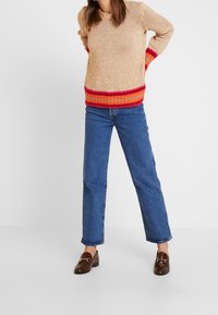Beige stickad tröja med röda och orange ränder vid nederkanten och ärmarna, tillsammans med blå raka jeans och bruna loafers.