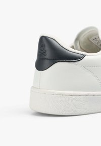 Adidași din piele albă cu accent din piele bleumarin la călcâi și logo cu craniu. Talpa din cauciuc texturat și căptușeala moale sunt vizibile. Design minimalist.