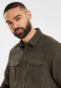 Donkergroene button-up shirt met een gestructureerde afwerking, voorzien van twee borstzakken met kleppen en bijpassende knopen. Gedetailleerde stiksels zichtbaar.