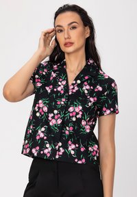 Chemise noire à manches courtes avec un motif floral rose et vert. Elle possède un col et des détails de boutons, avec une coupe relaxée et courte.