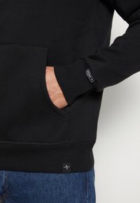 Schwarzer Sweatshirt mit einer Fronttasche, gerippten Bündchen und Saum. Auffälliges Logo-Tag an der unteren Seite über blauen Jeans. Weicher Stoff.