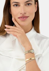Montre-bracelet en métal argenté et doré avec un cadran rectangulaire, portée avec un bracelet en or et un fin bracelet en argent sur une blouse claire.