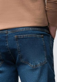 Vaqueros de mezclilla azul con una textura suave, con costuras en contraste de color naranja y dos bolsillos traseros. Diseño ajustado con cintura de altura media.