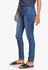 Kapeat siniset farkut, joiden pinta on sileä, viiden taskun muotoilu ja näkyvät tikit lahkeiden saumoissa. Yhdistetty mustiin litteisiin sandaaleihin.